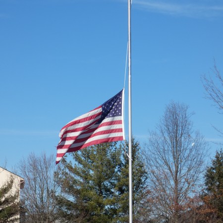 US flag flown at half-staff with trees and a blue sky in the background